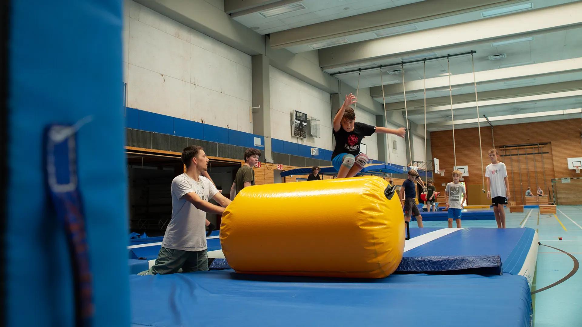 Kind springt begleitet von einem WEMOVE.-Trainer über eine Airtrack-Rolle